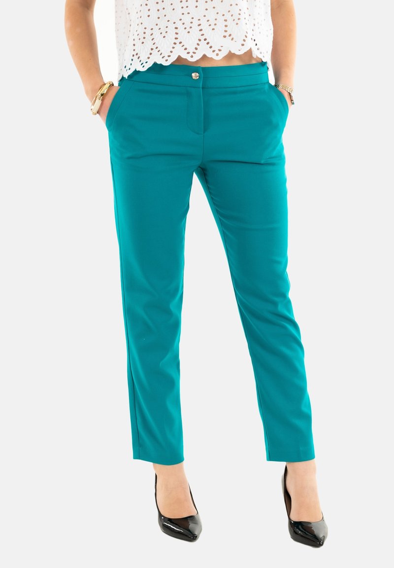 Pantalons ajustés teal avec poches latérales, une texture lisse, un fermoir et une fermeture éclair à l'avant, et un léger rétrécissement vers la cheville. Portés avec des talons noirs.