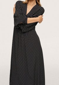 Černé šaty s puntíky a zavinovacím designem, dlouhými rukávy a sukní až na zem. Obsahují bílé puntíky a jemnou texturu látky.