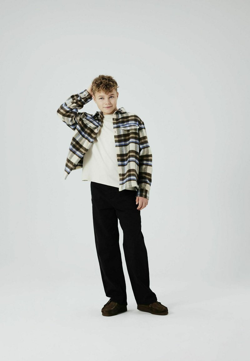 Camicia a flanella a quadri in verde, marrone, crema e blu, indossata sopra una maglietta a maniche lunghe crema; abbinata a pantaloni neri e scarpe marroni.