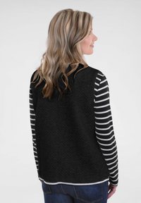 Zwarte geweven vest met hoge hals gedragen over een zwart-wit gestreepte longsleeve shirt. Achteraanzicht toont textuurdetails en zoom.