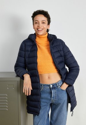 Personne souriante s'appuyant sur un casier gris, portant une doudoune marine, un pull orange à col roulé court et un jean bleu avec une main dans la poche.