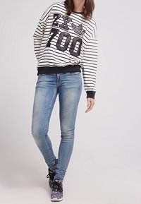 Černobílý pruhovaný svetr s velkým grafickým logem a číslem. Doplňují ho světle modré úzké džíny a vzorované černé tenisky.