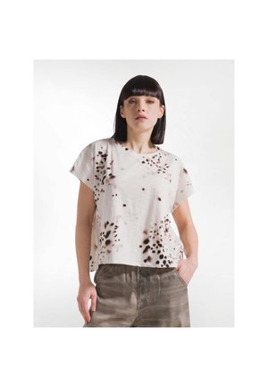 Femme aux cheveux raides et foncés portant une chemise blanche à manches courtes avec des motifs de taches foncées et un pantalon marron, une main dans la poche.