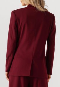 Burgundy tailored blazer with a notch lapel, single vent at the back, long sleeves, and smooth fabric texture.