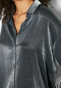 Glansig mörkgrå blus med krage, knäppning och textured tyg som skapar en subtil lyster.