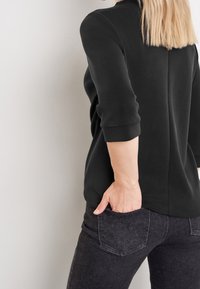 Schwarzes, weiches Stoffoberteil mit Dreiviertelärmeln, kombiniert mit dunkelblauen Jeans, die dezent sichtbare Gesäßtaschen aufweisen.