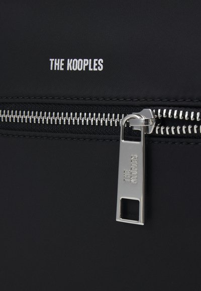 Melns neilona maisiņš ar rāvējslēdzēja detaļām, kas iekļauj sudraba vilkšanas elementu un šūtu apmalīti. "THE KOOPLES" logotips balta krāsā uzdrukāts virs rāvējslēdzēja.