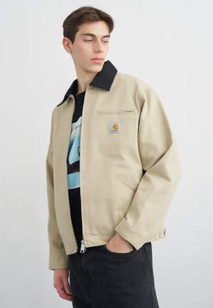 Νεαρός άνδρας που φορεί μπεζ μπουφάν Carhartt με μαύρο γιακά, μαύρο μπλουζάκι με γραφικό σχέδιο και σκούρο τζιν, που ποζάρει με το χέρι στην τσέπη, κοιτάζοντας προς τα αριστερά.