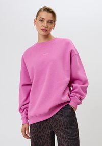 Felpa rosa oversize con scollatura rotonda e polsini a costine. Presenta la scritta bianca "OH APRIL" sul davanti. Indossata con pantaloni a stampa leopardata.