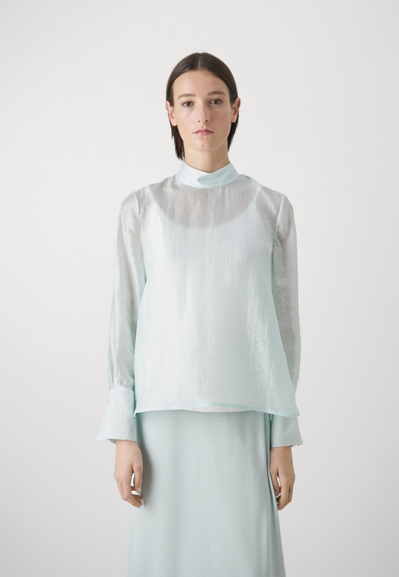 Top lungo con maniche trasparenti di un azzurro chiaro, con collo alto, texture liscia e vestibilità ampia; indossato sopra una gonna coordinata.