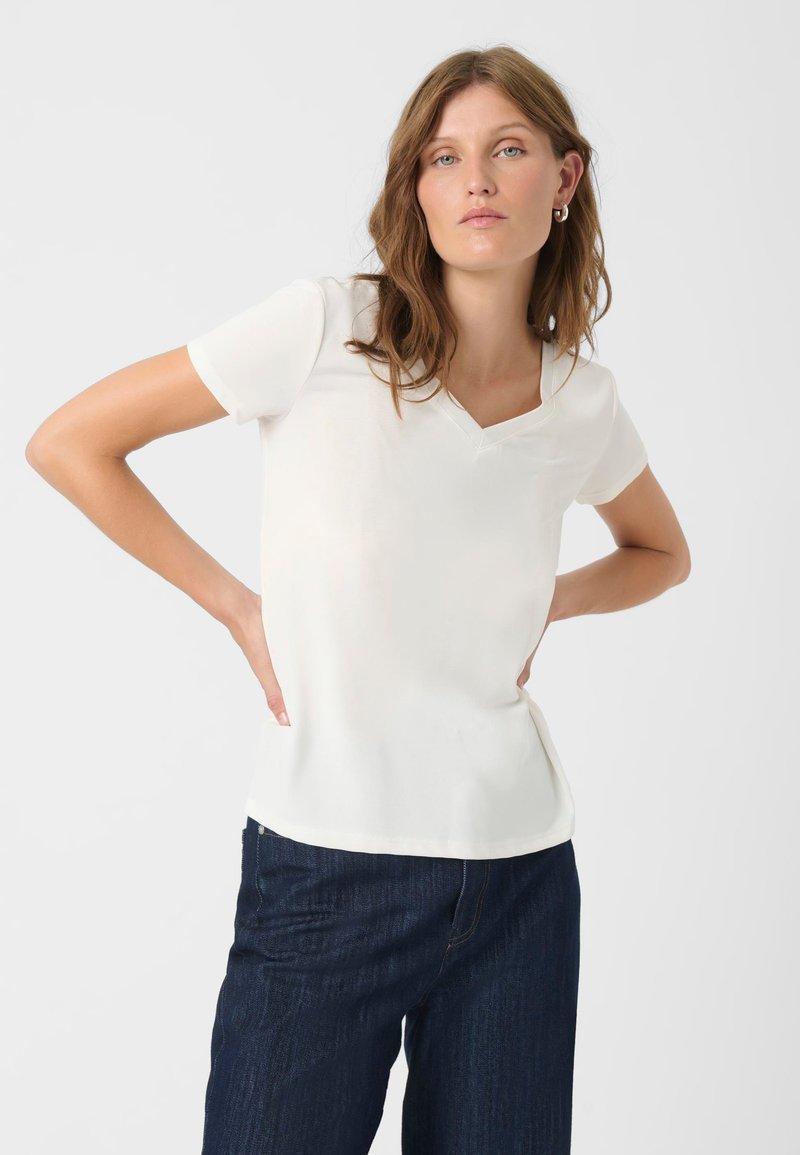 Haut blanc à manches courtes avec un col en V, texture douce, coupe ajustée et légères fentes sur les côtés, associé à un jean bleu foncé.