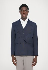 Tengerészeti, dupla soros blézer fekete gombokkal, csúcsos hajtókákkal és oldalsó zsebekkel, fehér galléros ing felett viselve, világosszürke nadrággal párosítva.