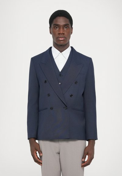 Blazer bleu marine à double boutonnage avec des boutons noirs, revers pointus et poches latérales, porté sur une chemise blanche à col, associé à un pantalon gris clair.