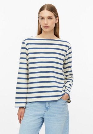 Vrouw met stijl bruin haar draagt een wit shirt met lange mouwen met horizontale blauwe strepen en lichtblauwe spijkerbroek, hand in zak.