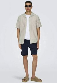 Chemise beige clair à manches courtes avec des boutons sur un t-shirt blanc, associée à des shorts bleu marine foncé et des sandales beiges. Le modèle porte des lunettes de soleil.