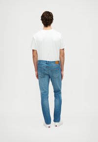 Kék farmernadrág, normál szabással, bőr folttal a hátsó derékrészen, sima fehér pólóval és fehér sportcipővel kombinálva.