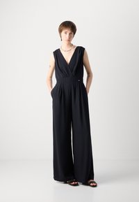 Marineblå uten ermer jumpsuit med dyp V-hals, vid ben-design og sidelommer. Jevn tekstur, minimale hardware-detaljer og skreddersydd passform.