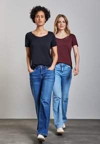 Due donne camminano in abbigliamento informale. La donna a sinistra indossa una maglietta nera a maniche corte e jeans blu. La donna a destra indossa una maglietta bordeaux e jeans azzurri più chiari.