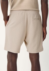 Bézs színű rövidnadrág halszálka mintával, rugalmas derékrésszel, oldalzsebekkel és egy hátsó zsebbel. A textúra szövöttnek és könnyűnek tűnik.