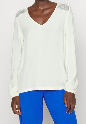 Weiße Bluse mit langen Ärmeln, V-Ausschnitt und Spitzenakzenten auf den Schultern. Verfügt über eine lockere Passform und strukturierten Stoff. Kombiniert mit blauen Hosen.