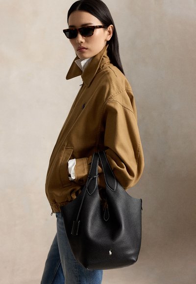 Persoon met een bruine jas en zonnebril houdt een grote zwarte leren shopper met wit logo vast tegen een effen achtergrond.