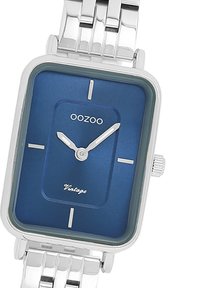 Montre-bracelet en métal argenté avec un cadran rectangulaire bleu, comportant des index horaires argentés, deux aiguilles et le nom de la marque "OOZOO" en haut.