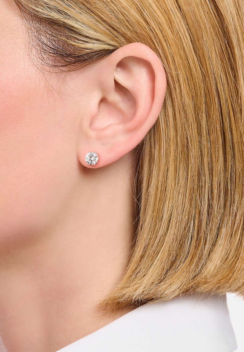 Runde Steckerohrringe aus klarem Kristall, gefasst in einem silberfarbenen Metall, die an dem Ohr eines Models mit glattem, blonden Haar platziert sind.