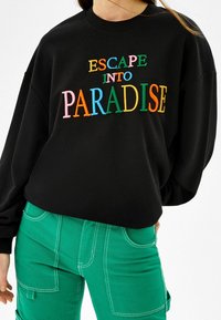 Svart sweatshirt med färgglad broderad text "ESCAPE INTO PARADISE" bärs med gröna byxor med vit sömnad och stora fickor.