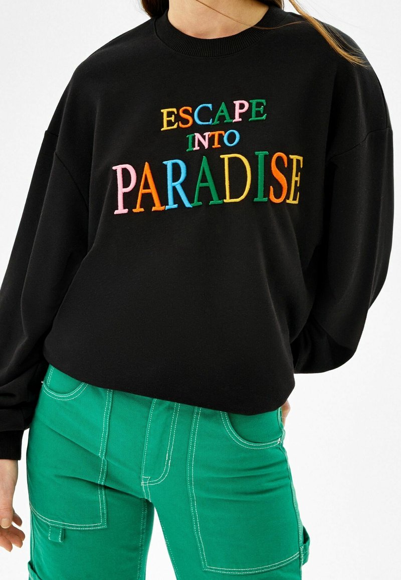 Svart sweatshirt med färgglad broderad text "ESCAPE INTO PARADISE" bärs med gröna byxor med vit sömnad och stora fickor.