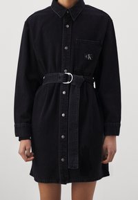 Zwarte denim jurk met knoopsluiting aan de voorkant, borstzak met logo-label, lange mouwen en een riem met een zilveren dubbele ringbuckle.
