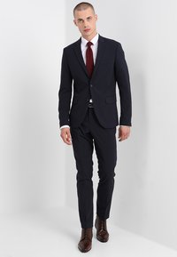 Marineblauer Anzug mit maßgeschneidertem Sakko und schmal geschnittenen Hosen, ergänzt durch ein weißes Hemd und eine weinrote Krawatte. Braune Lederschuhe runden den Look ab.