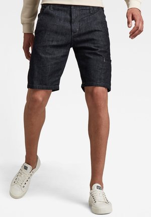 Dunkelblaue Jeansshorts mit regulärer Passform, umgeschlagenen Säumen und Fronttaschen, getragen mit weißen Sneakers. Glatte Textur und robuste Verarbeitung.