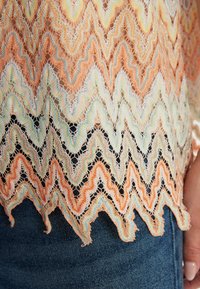 Kanten top met een zigzagpatroon in oranje, mintgroen en crèmekleuren, met een golvende zoom. G gedragen over donkerdenim. Translucente textuur.