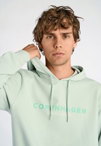 Sudadera verde menta con bolsillo canguro, capucha con cordón y "COPENHAGUE LANDBERGH" impreso en colores de contraste en la parte delantera.