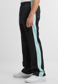 Crne sportske hlače s tirkiznim uzorkom sa strane, elastičnim pojasom s plavom vezicom i uparene s bijelim tenisicama.