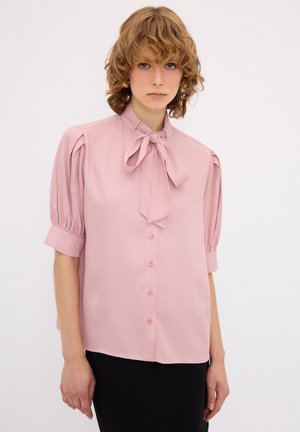 Blusa rosa realizzata in tessuto morbido, con colletto a fiocco, maniche corte a sbuffo e parte anteriore con bottoni abbinati.