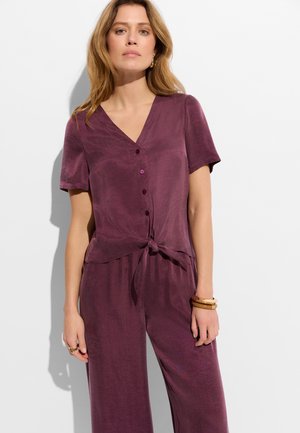 Kobieta ubrana w krótki rękaw śliwkowego topu zapinanego na guziki z wiązaniem z przodu i dopasowane szerokie spodnie, ozdobiona bransoletkami i pierścionkiem.