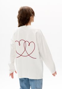 Weißes langärmeliges Sweatshirt mit lockerem Schnitt und einem roten, gestickten Herzdessin auf der Rückseite. Weiche Stoffstruktur und überschnittene Schultern.