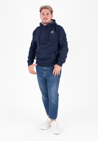 Felpa con cappuccio blu navy, dotata di tasca a marsupio, con lacci e un logo sul petto, abbinata a jeans blu sbiaditi e scarpe bianche.