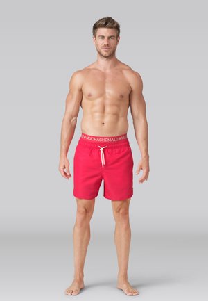 Svalnatý muž stojaci na boso proti jednoduchému pozadiu, oblečený v červených plavkách s bielym sťahovacím šnúrkou a nápisom "MUCHACHOMALO" na opasku.