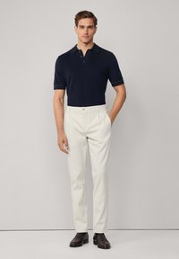 Muškarac stoji odjeven u mornarskoplavu majicu s kratkim rukavima, bijele krojene hlače i tamnosmeđe mokasine, nasuprot jednostavne svijetle pozadine.