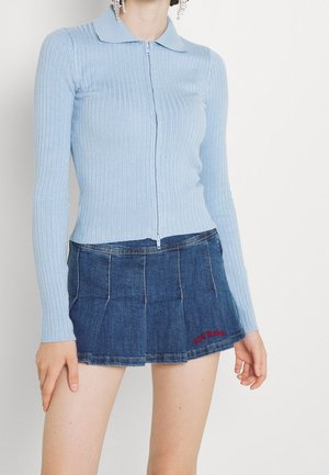 Woman wearing a light blue ribbed long-sleeve top with a collar and zipper, paired with a blue pleated denim mini skirt.