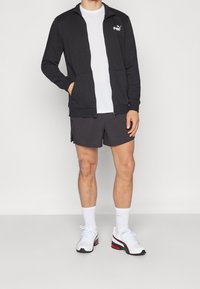 Giacca nera con zip e tasche, logo bianco; abbinata a pantaloni grigi e scarpe bianche con dettagli rossi; t-shirt bianca semplice sotto.