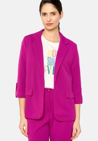 Blazer fucsia con rever a punta, due tasche anteriori e maniche a tre quarti, abbinato a una maglietta bianca con grafica e pantaloni coordinati.