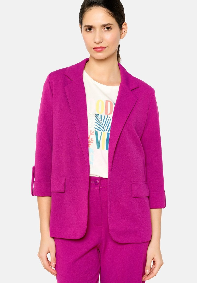 Blazer fucsia con rever a punta, due tasche anteriori e maniche a tre quarti, abbinato a una maglietta bianca con grafica e pantaloni coordinati.
