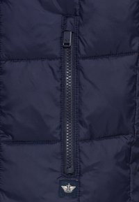 Marineblaue isolierte Jacke aus gestepptem Stoff mit einem zentralen Reißverschluss und einem Logopatch am unteren Rand. Glatte Textur und strapazierfähiges Material.