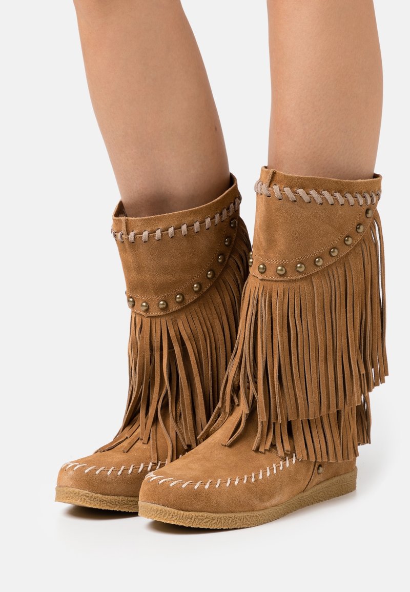 Bottines en suède beige avec des franges, ornées de clous métalliques, de surpiqûres contrastantes et d'une finition douce et texturée. Design à semelle plate.
