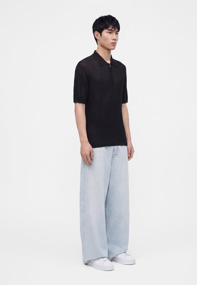 Jeune homme portant un polo noir côtelé à manches courtes, un jean bleu clair à jambes larges et des baskets blanches, debout devant un fond uni.