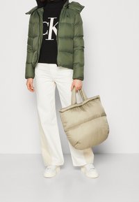 Zielona puchowa kurtka z kapturem, czarny dzianinowy sweter, białe szerokie spodnie oraz beżowa torba typu tote; cechują się gładką fakturą i przytulnym designem.