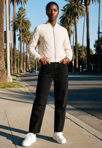 Giovane in piedi sul marciapiede vicino a palme, indossa una giacca trapuntata color crema, pantaloni neri e sneakers bianche con le mani in tasca.